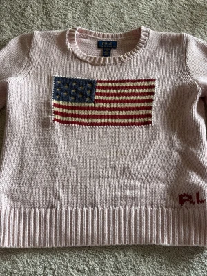 Ljusrosa stickad tröja Polo Ralph Lauren - Ljusrosa stickad tröja från Polo Ralph Lauren med amerikansk flagga i blått, rött och vitt på bröstet. Rund halsringning och ribbstickade muddar. Diskret RL-logo i rött vid nederkanten. Perfekt för dig som gillar klassisk och cool stil.