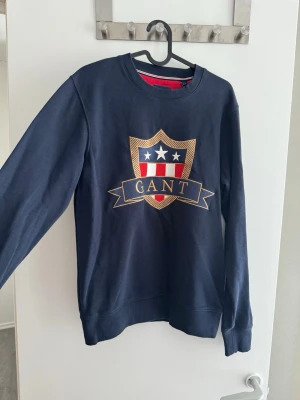 Mörkblå sweatshirt från GANT - Mörkblå sweatshirt från GANT med stort broderat emblem på bröstet i guld, rött, vitt och blått. Tröjan har rund halsringning och ribbade muddar vid ärmslut och nederkant. Perfekt för dig som gillar klassisk och sportig stil.