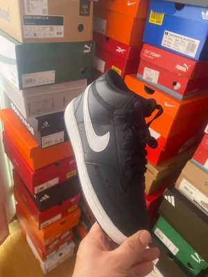 Svarta Nike sneakers med vit swoosh - Snygga svarta sneakers från Nike med klassisk vit swoosh på sidan. Skorna har en låg profil och är tillverkade i läder med perforeringar på tån för extra ventilation. Perfekt för dig som vill ha en stilren och tidlös look.