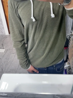 Olivgrön hoodie med vita snören - Snygg olivgrön hoodie med vita dragsnören i huvan. Hoodien har en avslappnad passform och långa ärmar, perfekt för en chill och casual stil. Tillverkad i mjukt material som känns skönt mot huden. Passar perfekt till jeans eller joggers.