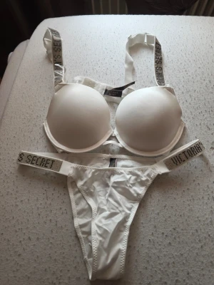 Vitt set från Victoria's Secret - Vitt underklädesset från Victoria's Secret med vadderad bh och matchande trosa. Bh:n har breda axelband med glittriga detaljer och text, och trosan har samma stil på banden. Materialet är mjukt och stretchigt för en skön känsla.