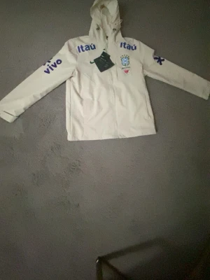 Vit Brasilien vindjacka med huva - Snygg vit vindjacka från Nike med Brasilien-logga och blåa sponsortryck på bröst och ärmar. Jackan har huva, dragkedja framtill och sportig look. Perfekt för dig som gillar fotboll och 