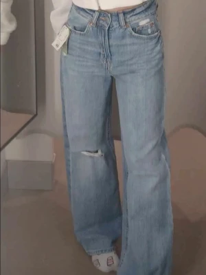 Vida ljusblå jeans med slitning - Säljer ett par trendiga ljusblå jeans med hög midja och vida ben. Jeansen har en liten slitning på ena knät och klassiska fem fickor. Perfekta för en avslappnad och cool stil. Materialet är denim i bomull.
