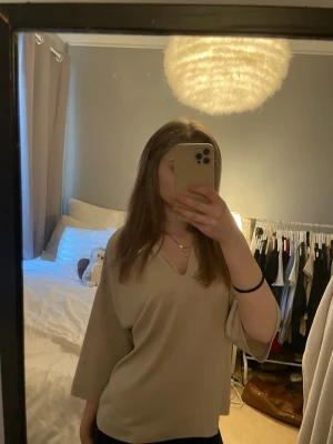Beige v-ringad blus med vida ärmar - Snygg beige blus med v-ringning och trekvartsvida ärmar. Blusen har en avslappnad passform och är tillverkad i ett mjukt, tunt tyg som faller fint. Perfekt att styla med jeans eller kjol för en clean look.