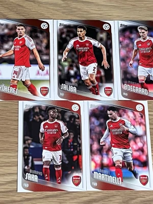 Arsenal Topps fotbollssamlarbilder - Fem samlarkort från Topps med Arsenalspelare: Gyökeres, Saliba, Ødegaard, Saka och Martinelli. Korten har färgstarka bilder på spelarna i rödvita matchställ och Arsenal-logga. Baksidan innehåller spelarinformation och statistik. Perfekt för fotbollsfans och samlare.