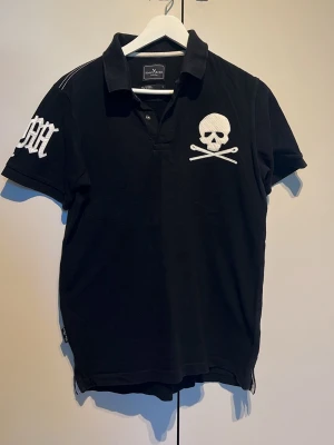 Svart t-shirt med dödskalle från Marc Ecko Cut & Sew - Cool svart t-shirt med vit dödskalle och korslagda ben på bröstet samt broderad detalj på ärmen. Klassisk krage och korta ärmar. Tillverkad i mjuk bomull för skön känsla och snygg passform.