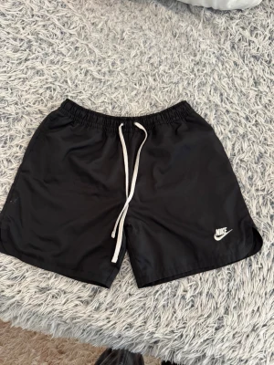 Svarta Nike shorts med snörning - Snygga svarta shorts från Nike med vit snörning i midjan och diskret Nike-logga på benet. Shortsen har elastisk midja och en praktisk bakficka med lock. Perfekta för sommarens alla aktiviteter.
