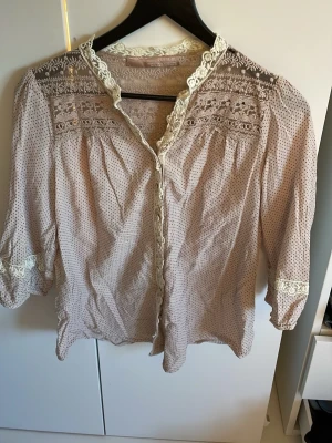 Beige spetsblus med trekvartsärm - Romantisk beige blus med små prickar och spetsdetaljer vid axlar, ärmslut och krage. Blusen har knäppning framtill och trekvartsärmar. Tillverkad i ett lätt och luftigt tyg, perfekt för en söt och vintageinspirerad look. Se tredje bilden för ett litet hål 