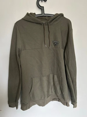 Olivgrön hoodie från River Island - Säljer en olivgrön hoodie från River Island i ribbad bomullsmix. Hoodien har huva med dragsko, magficka och en liten svart patch med logga på bröstet. Perfekt för en avslappnad och cool stil.