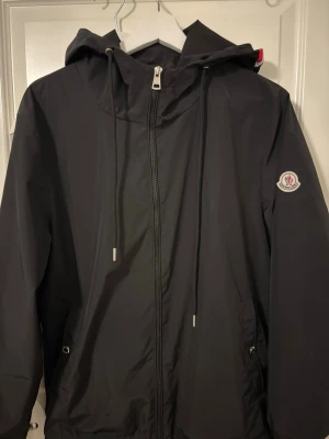 Svart Moncler vindjacka  - Svart vindjacka från Moncler med dragkedja framtill, justerbar huva med snören och logotyp på vänster ärm. Jackan har två fickor med dragkedja och är tillverkad i ett lätt, vattenavvisande material som passar perfekt för blåsiga dagar.