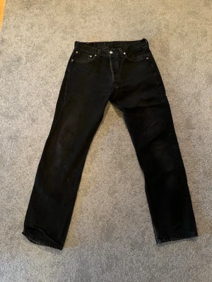 Svarta Levi's 501 - Säljer ett par klassiska svarta Levi's jeans med straight fit och femficksdesign. Jeansen har knappgylf och den ikoniska Levi's-lappen på bakfickan. Materialet är slitstark denim. Storlek W33 L32. Hör av er vid fler funderingar