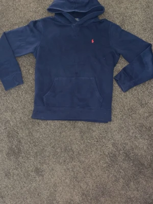 Mörkblå hoodie från Polo Ralph Lauren - Mörkblå hoodie från Polo Ralph Lauren med klassisk huva och känguruficka. Liten röd broderad logga på bröstet. Mjuk bomullskänsla och ribbade muddar vid ärmslut och nederkant. Perfekt för en avslappnad och stilren look.