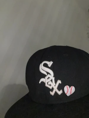Svart New Era White Sox keps - Snygg svart keps från New Era 59FIFTY med White Sox-logga i vitt framtill och ett broderat hjärta som ser ut som en baseboll. Klassisk rak skärm och broderade ventilationshål. Perfekt för dig som gillar sportig streetstyle.