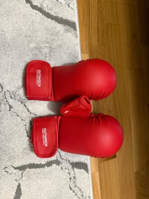 Röda kampsportshandskar Outshock - Säljer ett par röda kampsportshandskar från Outshock. Handskarna har kardborreband vid handleden för enkel justering och sitter skönt på handen. Tillverkade i syntetmaterial med vadderad ovansida för extra skydd. Perfekta för träning inom karate eller annan kampsport.