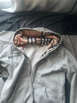Grå hoodie med Burberry-mönster - Säljer en grå hoodie med dragkedja från Burberry. Hoodien har klassiskt rutigt Burberry-mönster på insidan av huvan och längs insidan. Materialet är mjukt och bekvämt, perfekt för chill dagar. Stora fickor framtill och snören i huvan.