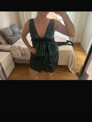 Mörkgrön omlottklänning med volang - Snygg mörkgrön kortklänning med djup v-ringning och omlottdetalj i midjan. Klänningen har volang nedtill och markerad midja med knytband. Perfekt för dig som vill sticka ut med en elegant och trendig look.