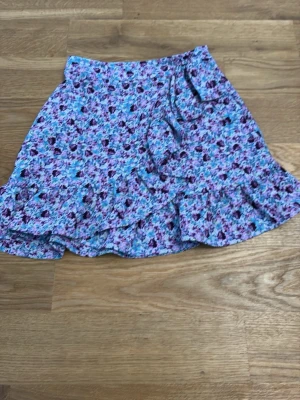 Blommig volangkjol i blått och lila - Söt kortkjol med blommigt mönster i blåa och lila nyanser. Kjolen har volangdetaljer längs nederkanten som ger en lekfull look. Perfekt för dig som gillar färg och mönster. Materialet känns lätt och luftigt.