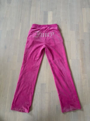 Rosa velourbyxor från Juicy Couture - Säljer ett par ikoniska rosa mjukisbyxor från Juicy Couture i velour. Byxorna har raka ben, resår och dragsko i midjan samt fickor fram. På baksidan finns Juicy-loggan i glittriga stenar. Perfekta för en chill och trendig look. Storlek S men funkar även på M
