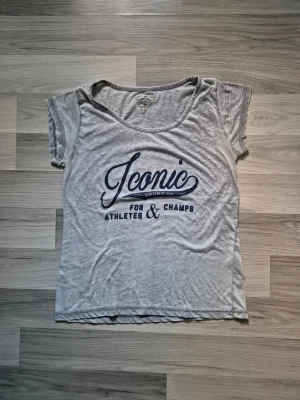 Grå Iconic t-shirt med tryck - Cool grå t-shirt från Iconic Sports Co. med blått tryck på framsidan som säger 'Iconic for athletes & champs'. T-shirten har en avslappnad passform, rund hals och korta ärmar. Perfekt för dig som gillar sportiga och casual vibes.