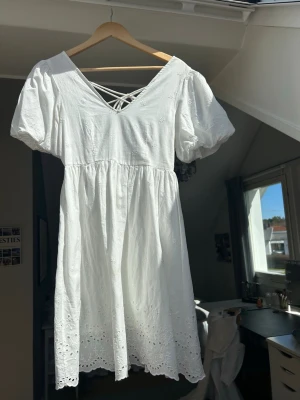 Vit kortklänning med snörning - Söt vit kortklänning med puffärmar och snörning i ryggen. Klänningen har broderade små blommor och v-ringad front. Materialet är bomull och nederkanten har fina spetsdetaljer. Perfekt för sommarens alla tillfällen eller till skolavslutning eller student! Endast använd en gång under skolavslutningen. 