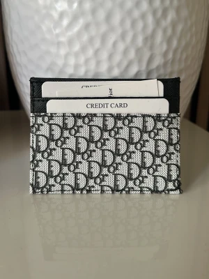  grå mönstrad kortfodral - . Fodralet har flera fack för kort och är tillverkat i canvas med detaljer i svart läder. Perfekt för att hålla dina kort organiserade och stilrena.
