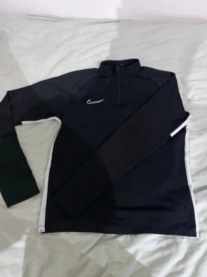 Svart Nike Dri-FIT träningströja - Svart långärmad träningströja från Nike med vit logga på bröstet och vita detaljer längs sidorna. Tröjan har halv dragkedja och är tillverkad i Dri-FIT material som transporterar bort fukt. Perfekt för sport och träning.
