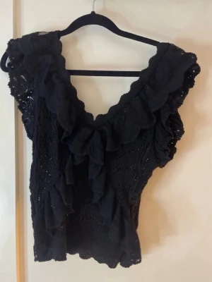 Svart spetsblus med volang - Säljer en svart blus i spets med djup v-ringning och stora volangdetaljer framtill. Toppen är ärmlös och har en feminin, elegant look som passar perfekt till jeans eller kjol. Perfekt för dig som vill sticka ut lite extra.