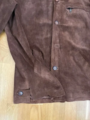 Brun skinnjacka från Molltan - Säljer en brun skinnjacka från Molltan med klassisk knappstängning och två snedställda fickor framtill. Jackan har en slät design i mjukt skinn och en innerficka. Perfekt för dig som vill ha en stilren och tidlös look.