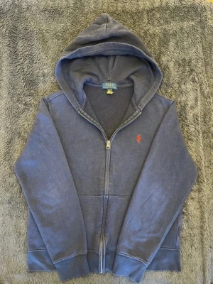 Blå zip hoodie från Polo Ralph Lauren - Mörkblå hoodie med dragkedja från Polo Ralph Lauren. Klassisk design med huva, känguruficka och röd broderad logga på bröstet. Tillverkad i mjukt bomullsmaterial som är perfekt för chill dagar. Enkel och stilren look som funkar till det mesta.