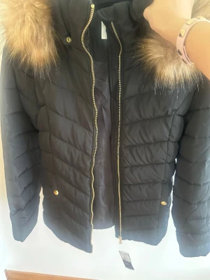 Svart pufferjacka med pälsdetalj - Svart pufferjacka med guldiga dragkedjor och knappar. Jackan har en stor huva med fluffig brun fuskpäls runt kanten. Quiltad design och två fickor framtill. Perfekt för kalla vinterdagar och riktigt snygg att bära utomhus. Helt ny med prislapp.