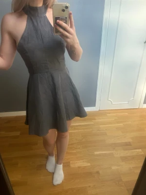 Grå kort klänning med snörning - Supersnygg grå kort klänning med hög hals och öppen rygg. Klänningen har snörningsdetaljer i ryggen som ger en unik look och en figurnära passform upptill med utsvängd kjol. Klänningen är väldigt mjuk.