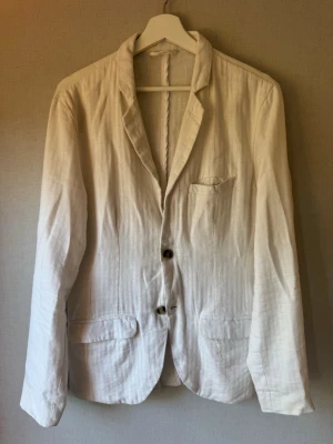 Ljusbeige kavaj från Lindbergh - Stilren ljusbeige kavaj från Lindbergh White i slim fit-modell. Tillverkad i en mix av bomull och linne för en luftig känsla. Tvåknäppt med klassisk krage, bröstficka och två framfickor. Perfekt för en fräsch och avslappnad look.