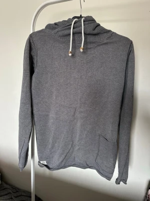 Grå hoodie med snörning och ficka - En stilren grå hoodie med huva och vita snören med trädetaljer. Hoodien har en praktisk ficka på framsidan och är tillverkad i mjukt material. Insidan av huvan är ljusare och ger en snygg kontrast. Perfekt för dig som gillar enkel och clean stil.