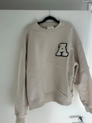 Beige sweatshirt från Axel Arigato - Säljer en beige  oversize sweatshirt från Axel Arigato med ett stort A-märke på bröstet. Tröjan har rund hals, ribbade muddar och är gjord i mjukt bomullsmaterial. Perfekt för dig som gillar stilrena och trendiga plagg med en sportig touch.