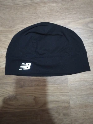 Svart mössa från New Balance - En svart mössa från New Balance med logotyp i vitt på framsidan. Mössan är tillverkad i ett stretchigt och mjukt material, perfekt för sport och vardag. Enkel design med sömmar som ger en snygg passform.