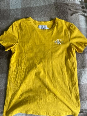 Gul t-shirt från Calvin Klein Jeans - Säljer en snygg gul t-shirt från Calvin Klein Jeans med klassiskt CK-logo broderat på bröstet. T-shirten har rund hals och korta ärmar, perfekt för dig som vill ha en enkel men trendig look. Materialet är mjuk bomull som känns skönt mot huden.
