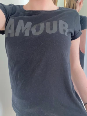 Svart t-shirt med AMOUR-tryck - Snygg svart t-shirt med stort tryck 'AMOUR' över bröstet. Klassisk rund halsringning och korta ärmar. Perfekt till jeans eller shorts för en avslappnad look. Materialet känns mjukt och bekvämt, troligtvis bomull.