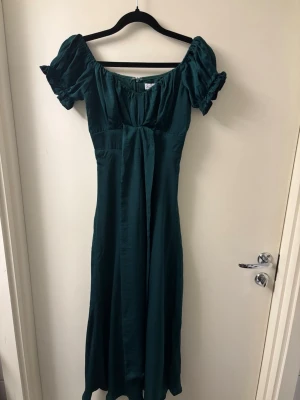 Mörkgrön omlottklänning med rosett - Supersöt mörkgrön klänning med omlottdetalj och rosett i ryggen. Klänningen har korta puffärmar och en smickrande v-ringning. Perfekt för dig som vill ha en feminin och trendig look. Säljer då den är för liten för mig. Aldrig använd, nyskick. OBS lånade bilder då den inte passar mig. Kontakta mig vid frågor. 