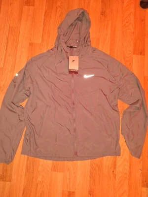 Nike vindjacka med huva grå S - Nike vindjacka i grått med hel dragkedja och huva. Jackan har reflekterande logga på bröstet och är tillverkad i lätt, vindtätt material. Perfekt för löpning eller promenader när det blåser. Två sidofickor och justerbar huva.