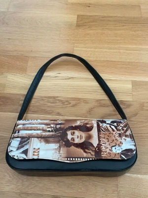 Svart handväska med vintage-tryck - Cool svart handväska med vintage-inspirerat tryck i brunt och vitt på framsidan. Trycket visar retro foton och text, vilket ger väskan en unik och konstnärlig vibe. Bredd-28 cm Höjd-13 cm, Djup 5,5 cm.