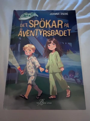 Det spökar på äventyrsbadet - En spännande bok av Joanna André med titeln 'Det spökar på äventyrsbadet'. Omslaget är färgglatt med illustrationer av två barn i pyjamas på ett äventyrsbad, och en mystisk bläckfisk i bakgrunden. Boken har mjukt omslag och riktar sig till unga läsare.