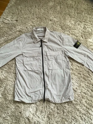Beige overshirt från Stone Island - Säljer en beige skjorta från Stone Island med svart dragkedja framtill och två stora bröstfickor. Skjortan har lång ärm och klassisk krage samt den ikoniska loggan på vänster ärm. Perfekt för en avslappnad men stilren look. Storlek M