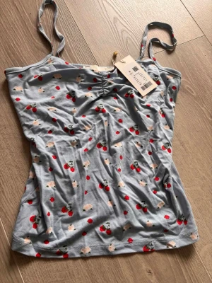 Ljusblå linnetopp med körsbärsmönster - Supersöt ljusblå linnetopp med tunna axelband och körsbärsmönster i rött och grönt samt små vita blommor. Toppen har en lätt rynkning vid bysten och är perfekt för varma dagar. Materialet känns mjukt och stretchigt.