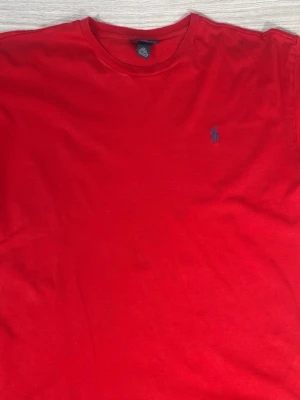Röd t-shirt från Polo Ralph Lauren - Klassisk röd t-shirt från Polo Ralph Lauren med rund halsringning och den ikoniska lilla blå loggan broderad på bröstet. Tillverkad i mjuk bomull och har en normal passform. Perfekt till jeans eller shorts för en clean look.