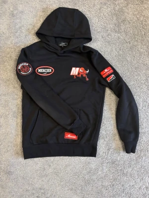 Svart hoodie från Mercier med patches - Svart hoodie från Mercier med röda och vita broderade patches på bröstet, ärmarna och en stor Mercier-logga på ryggen. Hoodien har huva och känguruficka framtill. Materialet ser ut att vara mjuk bomullsblandning. Snygga detaljer och sportig vibe.
