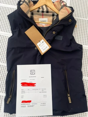 Marinblå dunväst från Burberry - Snygg marinblå dunväst från Burberry med klassiskt rutigt foder och huva. Västen har två framfickor med guldfärgade dragkedjor och detaljer i metall. Perfekt för lager på lager och en stilren look.