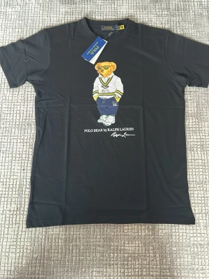 Svart t-shirt med Polo Bear från Ralph Lauren - Cool svart t-shirt från Ralph Lauren med Polo Bear-motiv på framsidan. Björnen har på sig en vit tröja med RLTC-tryck och blå byxor. T-shirten har rund hals och korta ärmar, perfekt för dig som gillar klassiska och trendiga prints.