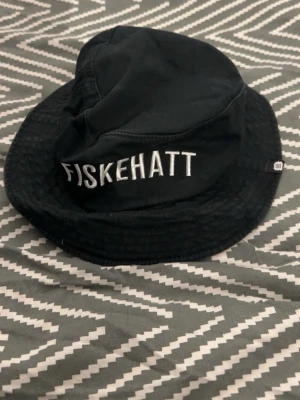 Svart fiskehatt med broderad text ODZ - Säljer en svart fiskehatt i tyg med vit broderad text 'FISKEHATT' framtill. Klassisk bucket hat-modell som ger en avslappnad och trendig look. Perfekt accessoar för dig som vill sticka ut med humor och stil.