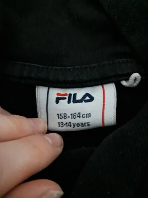 Svart hoodie från FILA med logga - Svart hoodie från FILA med stor vit och röd logga på bröstet. Klassisk modell med huva och magficka, ribbade muddar vid ärmslut och nederkant. Perfekt för en avslappnad och sportig stil.