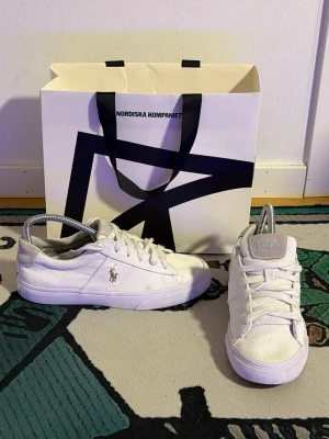 Vita Polo Ralph Lauren sneakers - Snygga vita sneakers från Polo Ralph Lauren med klassisk logga på sidan och POLO-text på hälen. Skorna har lågt skaft, vita snören och är tillverkade i canvas med mockadetalj på hälen. Perfekt för dig som gillar stilrena och trendiga sneakers.
